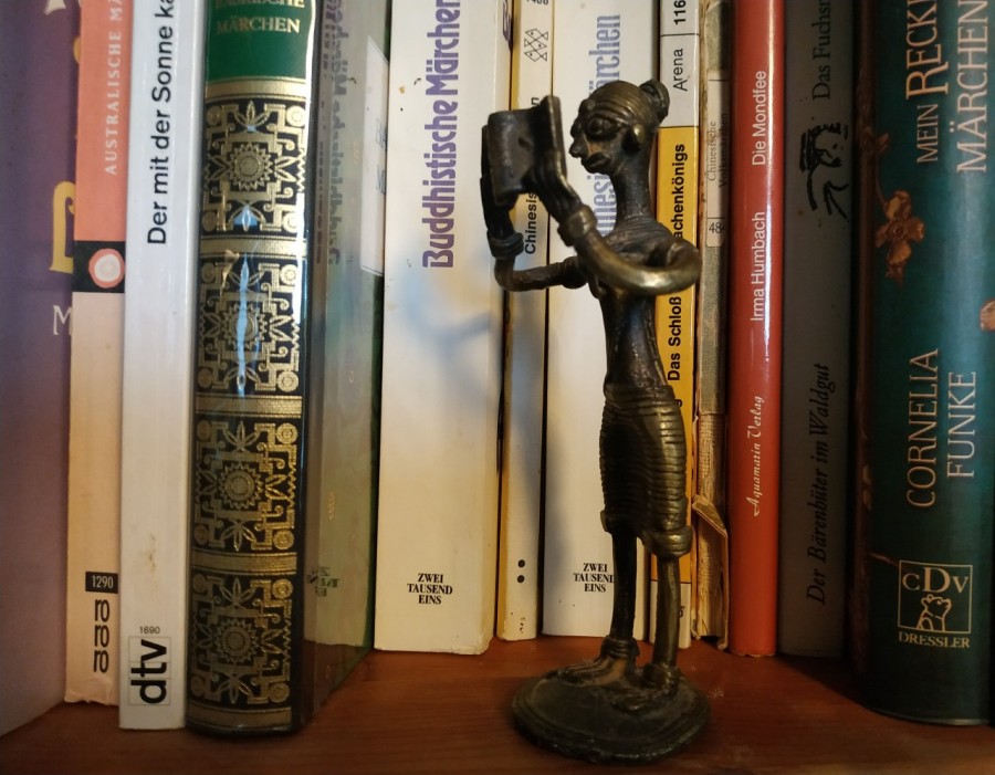 Im Regalfach vor Märchenbüchern steht die Messingfigur einer lesenden Frau mit Brille