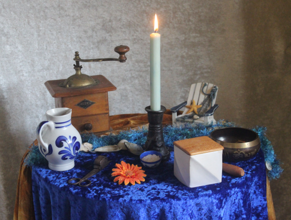 Auf einem runden Tisch, der mit einem blauen Pannésamttuch bedeckt ist stehen im Kreis von links nach rechts, ein Krug aus Steingut mit blauem Muster, eine mechanische Kaffeemühle aus Holz, ein Kerzenständer aus Bronze mit einer brennenden hellblauen Kerze, ein maritim dekorierter weißer Schauekelstuhl, eine Klangschale, ein würfelförmiger weißer Behälter mit einem Deckel aus unbehandeltem hellem Holz, eine orngefarbene Platikblüter und eine Lichtputzschere