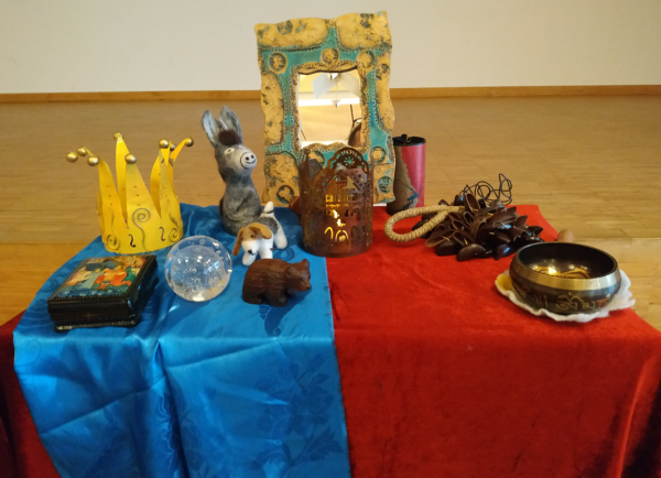 Auf einem rechteckigen Tisch liegen ein rotes Pannésamttuch, auf der linken Seite bedeckt von einem blauen mongolischen Seidentuch (Haddak). Darauf stehen von links nach rechts in der jinteren Reihe eine gelbe Blechkrone mit goldenen Kugeln an den Spitzen, ein Filzesel, ein Spiegel mit einem Rand aus naturfarbener Keramik mit türkisfarbenem Muster, mein rotes Zafir-Klangspiel Sunray, meine Cabona-Rassel und meine Klangschale aus Bhutan, in der zweiten Reihe das russiche Kästchen für meine Visitenkarten, die transparente Glaskugel mit den Lufteinschlässen, ein Filzhund und ein Bär aus Holz. Vor dem Spiegel steht ein Teelichthalter aus braun eloxiertem Blech, der aus 5 Giebelseiten aötmodischer Häuser besteht.