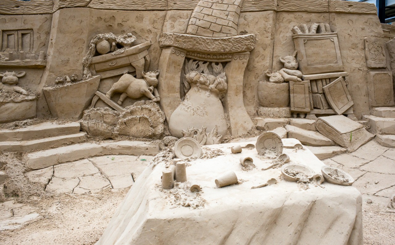 Die Sandskulpturen zeigen drei dicke Geißlein, die sich verstecken und dabei das Geschirr auf dem Tisch im Vordergrund umgeworfen haben