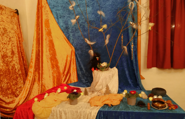 Tisch mit orangefarbenen, blauen und roten Stoffen, darauf Zweige mit Federn, kleine Töpfe mit Pflanzen und verschiedene Dekorationsgegenstände