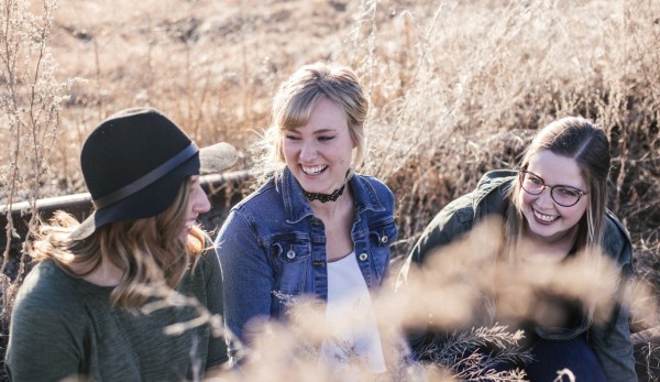 Drei Frauen sitzen im Freien auf trockenem Gras, die erste trägt einen schwarzen Hut, die zweite eine Jeansjacke und die dritte dunkle Jacke