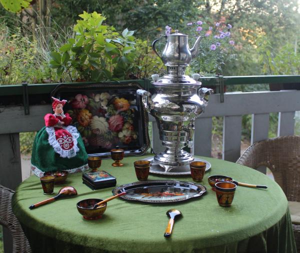 Großer silberner Samowar mit Teekanne auf grünem Tisch mit bemalten Holzschalen, Löffeln, Tablett und einer Puppe in traditioneller Tracht vor einem Balkon mit Pflanzen