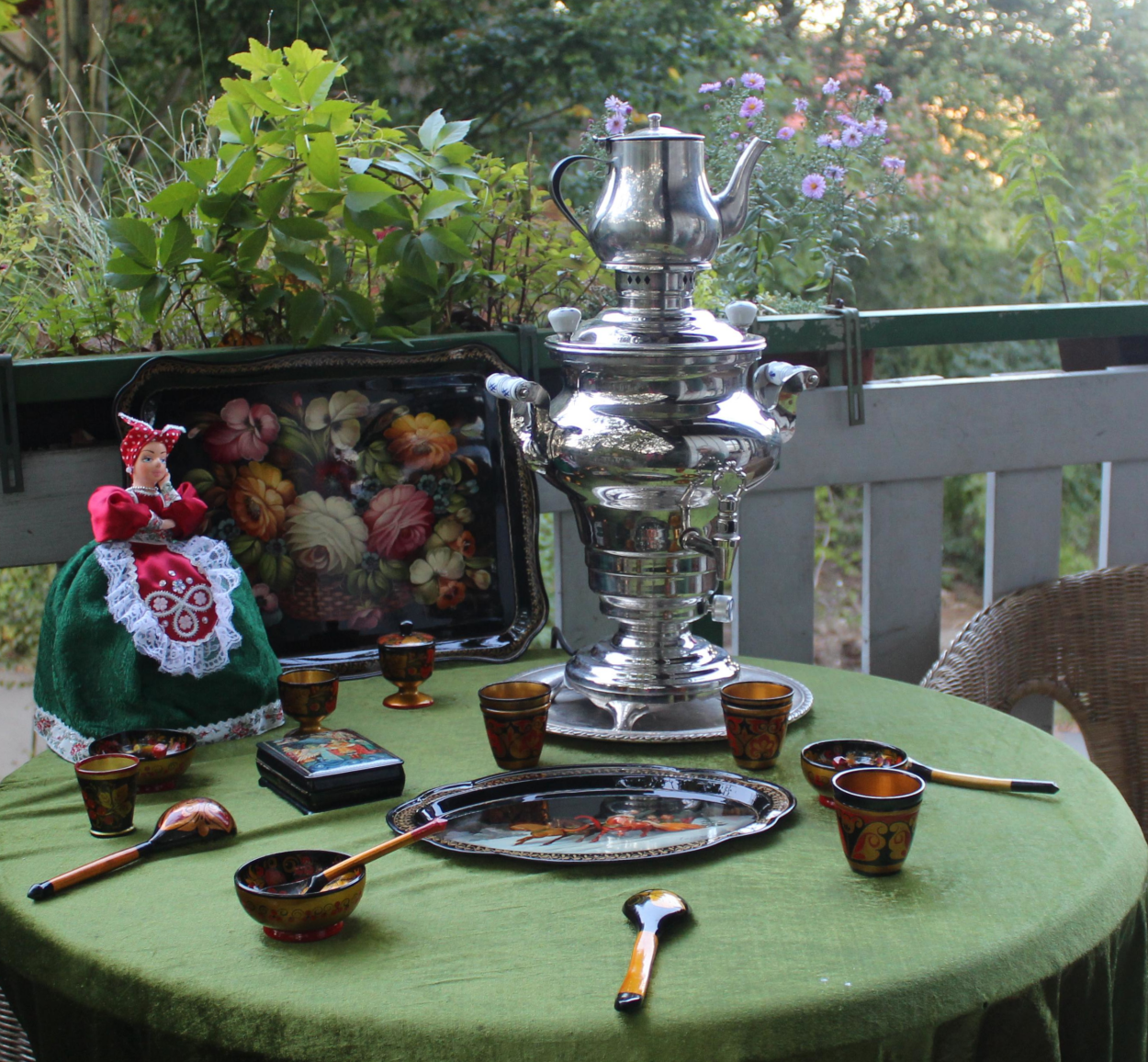 Großer silberner Samowar mit Teekanne auf grünem Tisch mit bemalten Holzschalen, Löffeln, Tablett und einer Puppe in traditioneller Tracht vor einem Balkon mit Pflanzen