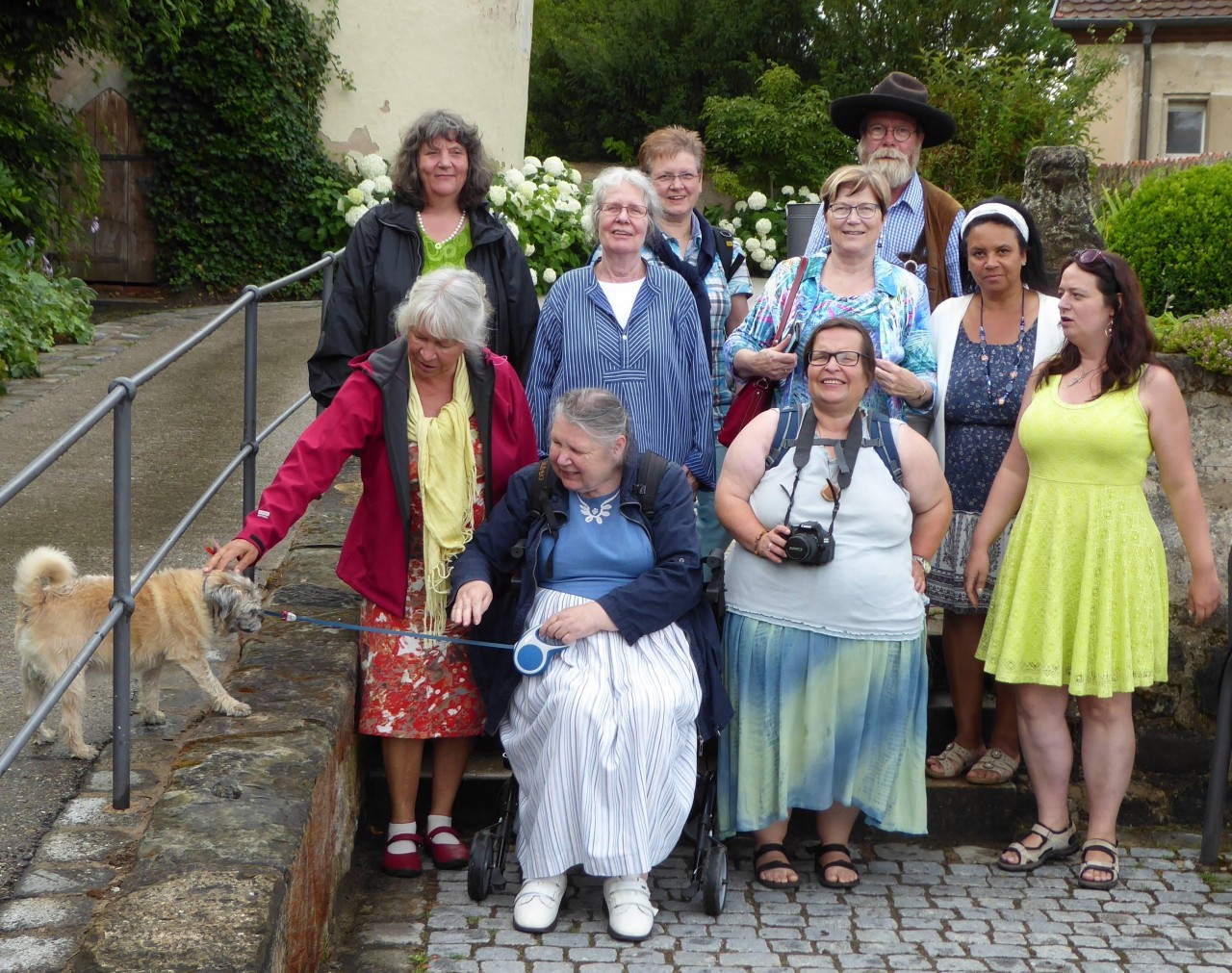Eine Gruppe von neun Frauen und einem Mann, darunter eine auf einem Rollator sitzend, steht auf gepflastertem Weg vor einem Haus, eine Person hält einen Hund an der Leine