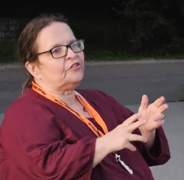 Ich trage ein dunkelrote Oberteil und ein orangefarbenes Schlüsselbändchen und gestikuliere mit beiden Händen vor dunklem Hintergrund