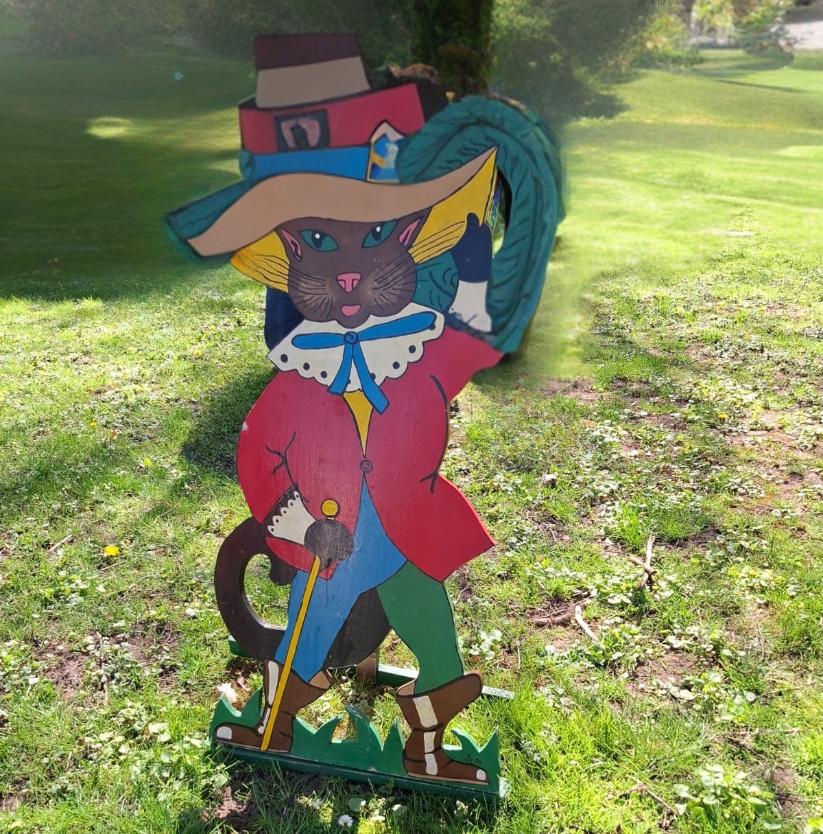 Holzfigur des gestiefelten Katers mit Hut, Feder, rotem Mantel und Stiefeln auf einer Wiese