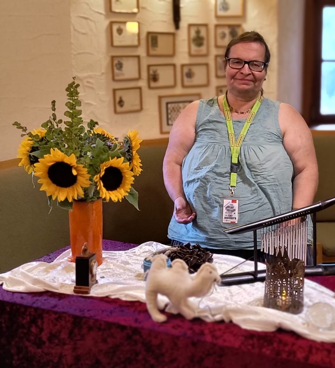 Bettina in ein  hellblaues ärmelloses Oberteil gekleidet steht hinter einem Tisch mit Sonnenblumenstrauß, weißem Stoff, weißem Filzkamel und Laterne in einem Raum mit Bilderwand